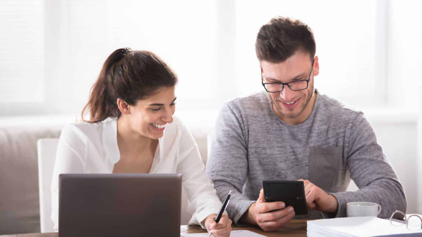 Happy Young Couple Calculating Bills At Home Using Calculator And Laptop