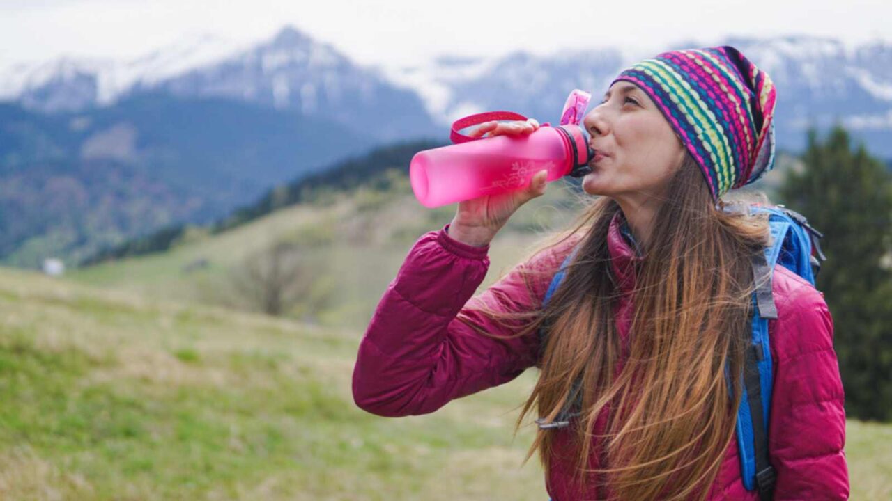 Woman hiker with backpack driking from reusable water plastic bottle outdoors in nature