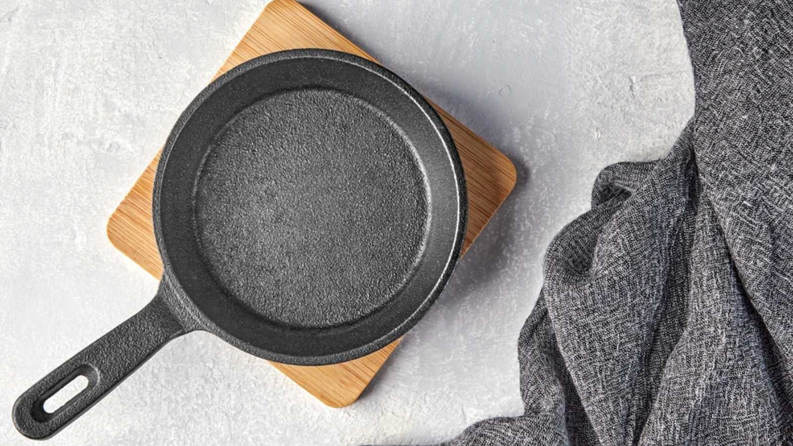 Black cast iron empty frying pan for one person on a wooden stand and a gray kitchen towel on a gray concrete background. Top view with copy space