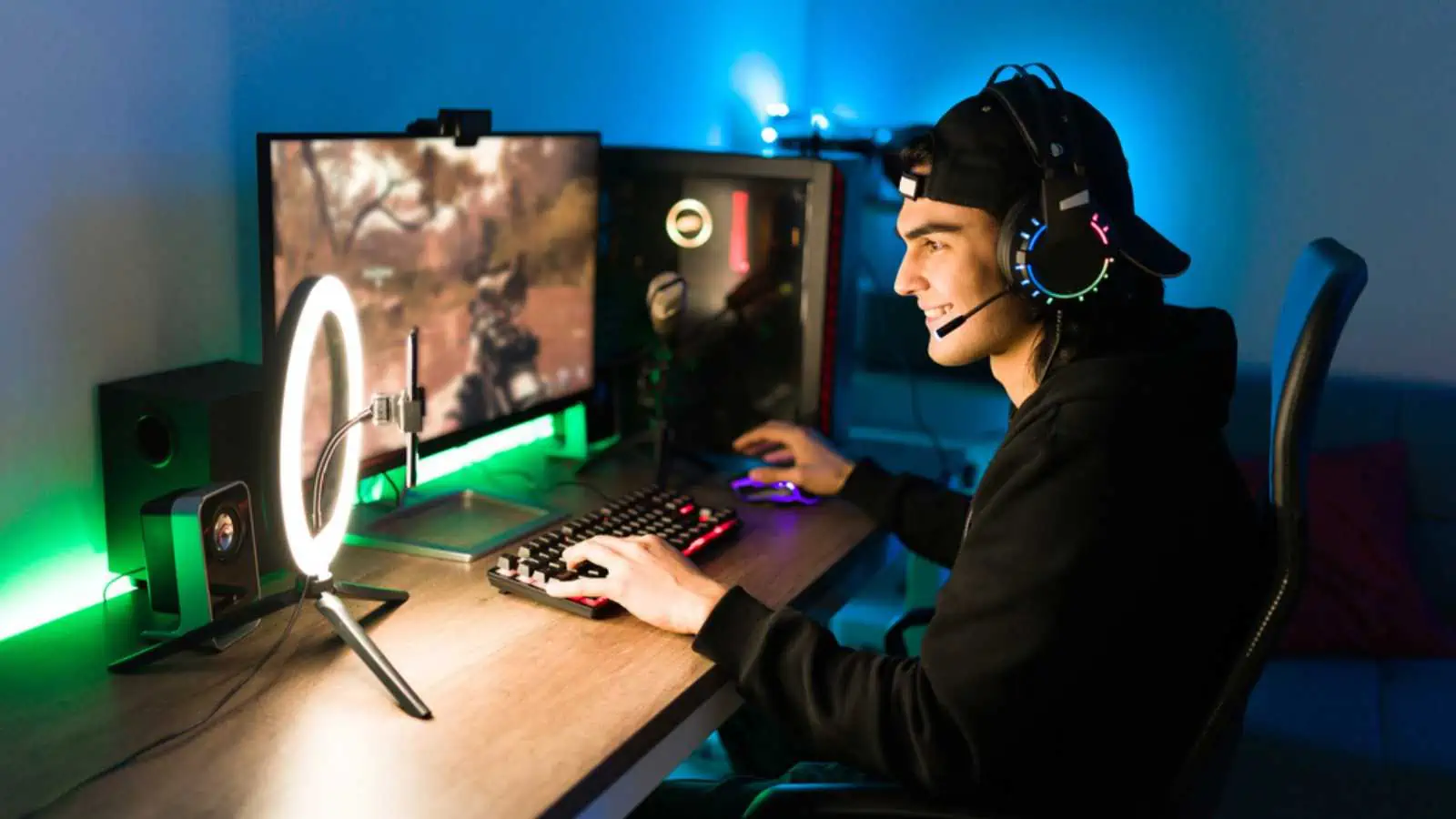Smiling young man live streaming his online video game using a smartphone and a ring light. Happy gamer ready to start playing in a gaming computer