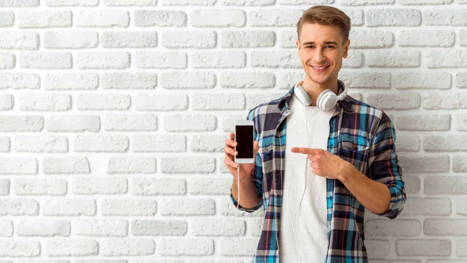 Young handsome man show the index finger at a blank black screen smartphone in casual style clothing against the backdrop of a brick wall. Close-up