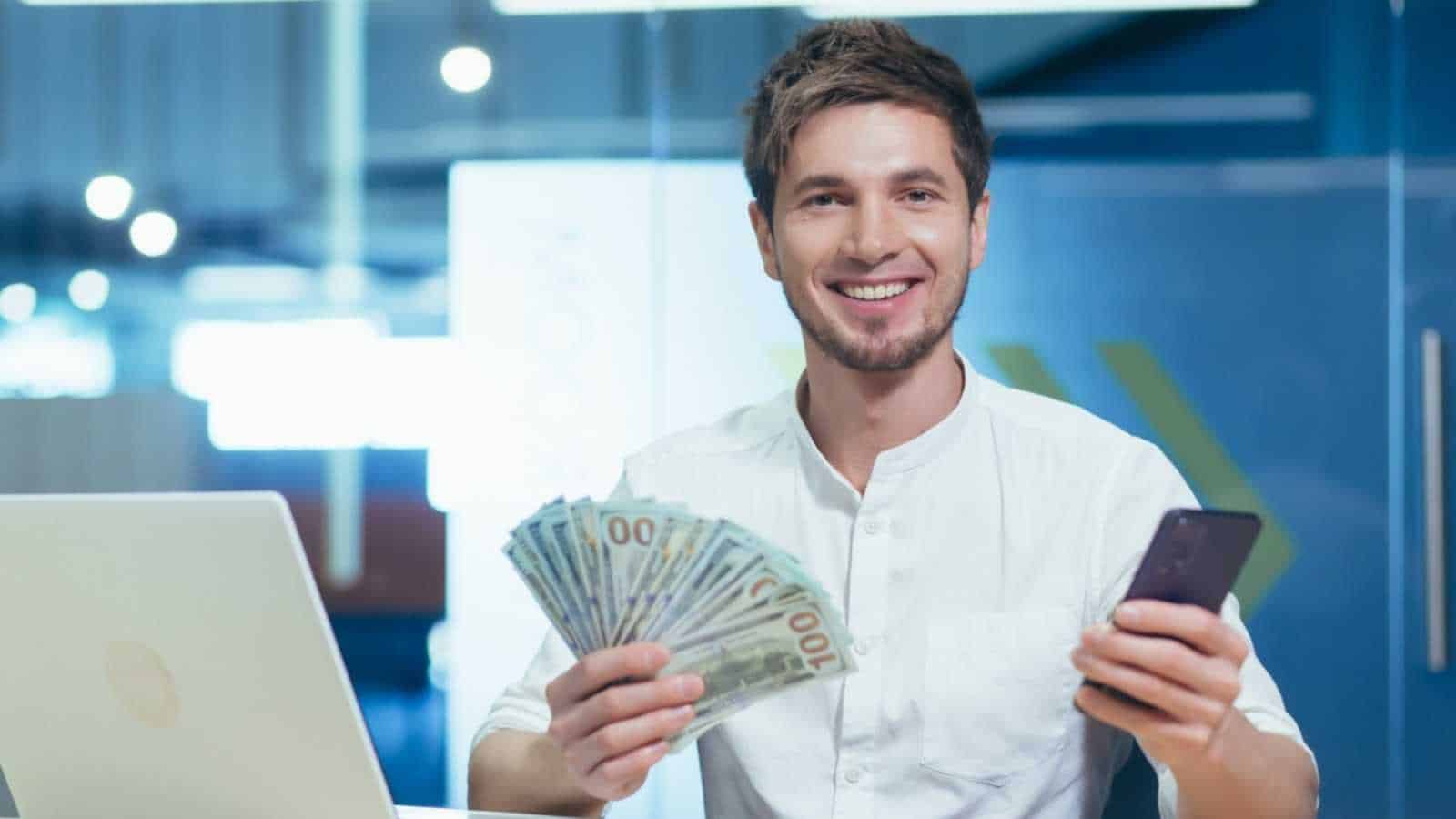 Happy and satisfied young handsome man. Businessman holding a wad of money and a mobile phone. Sits in the office at the desk, shows, brags. smiling