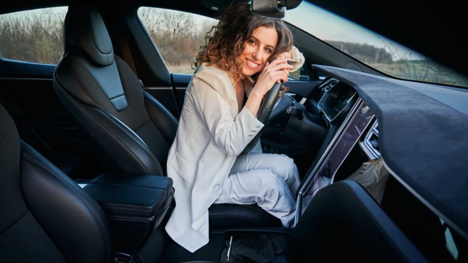 Portrait of beautiful curly lady inside electric car interior who putting arms on steering wheel. Smiling girl with curly hair adoring her automobile.