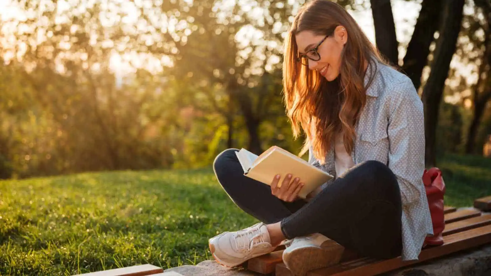 Side view of pleased brunette woman in eyeglasses sitting on bench and reading book in park