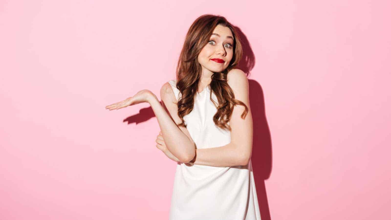 Portrait of a uncertain pretty woman shrugging shoulders isolated over pink background